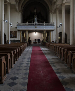 Photograph-of-an-empty-dimly-lit-church-with-wooden-pews-red-carpet-aisle-stone-columns-arched-ceiling-and-stained-glass-windows