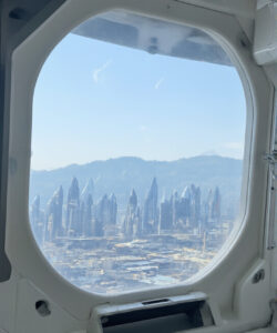Photograph-of-futuristic-cityscape-through-airplane-window-featuring-sleek-skyscrapers-and-distant-mountains-under-a-clear-blue-sky