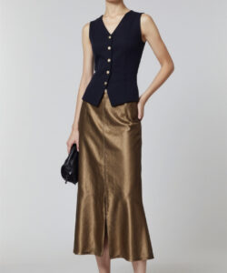 Photograph of slender asian woman in black vest gold skirt black sandals holding black bag standing against plain white background