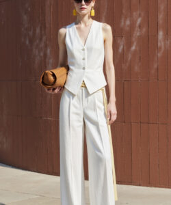 Photograph of slender white woman in white pantsuit holding brown hat against red textured wall wearing yellow earrings sunglasses