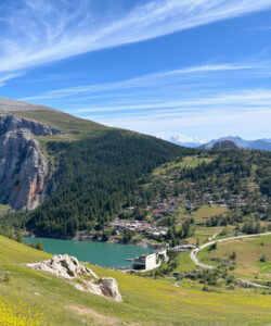 panoramic-photograph-of-a-mountainous-landscape-clear-blue-sky-scattered-clouds-greenery-yellow-wildflowers-rocky-cliffs-serene-lake-village