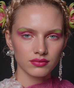 A Fair Skinned Woman With Braided Hair Vibrant Makeup And Pearl Earrings Against A