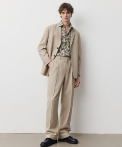 A Young Man In A Beige Suit Floral Shirt Black Shoes Against A Plain White Background