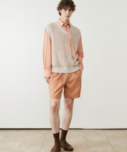 A Young Man With Curly Hair Wearing A A Knitted Vest Peach Shirt Tan Shorts And