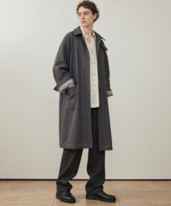 A Young White Man With Curly Hair Wearing Wearing A Long Dark Overcoat Beige Shirt And