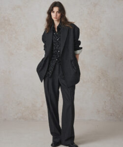 A Young Woman With Long Wavy Brown Hair Hair Wearing A Black Suit Over A Black