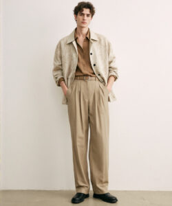 Curly Haired Man In A Beige Coat And Tan Pants Hands In Pockets Against A Plain