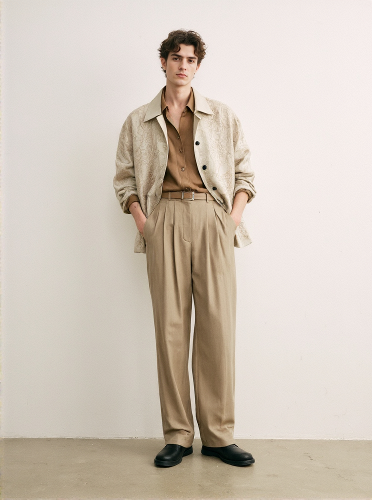 Curly Haired Man In A Beige Coat And Tan Pants Hands In Pockets Against A Plain