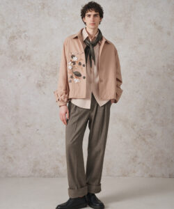 Curly Haired Man In A Beige Jacket Brown Pants Black Shoes Against A Textured