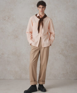 Curly Haired Man In A Peach Shirt Brown Tie Beige Trousers And Black Shoes Against A