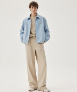 Curly Haired Man In Light Blue Shirt Shirt Beige Pants And Sneakers Standing Against