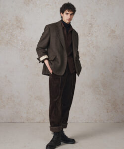 Curly Haired Young Man In A Brown Velvet Suit Black Boots Against A Textured