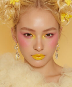 With Blonde Hair In Pigtails Wearing A Crown Yellow Makeup And Yellow Earrings Against
